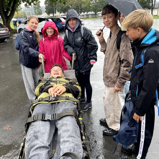 Eine Gruppe junger Personen versammelt sich um einen Jungen auf einer Trage, hält Regenschirme und trägt Regenjacken auf einem nassen Parkplatz.