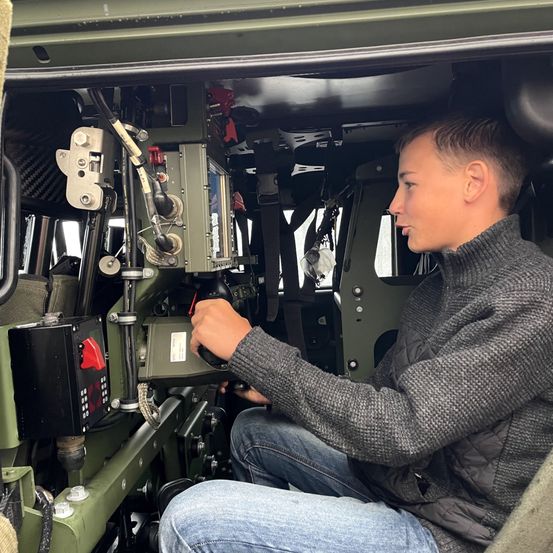 Ein junger Mann in einem grauen Pullover und Jeans sitzt in einem Militärfahrzeug und hält das Lenkrad. Verschiedene Steuerungen und ein Monitor sind im Innenraum des Fahrzeugs sichtbar.