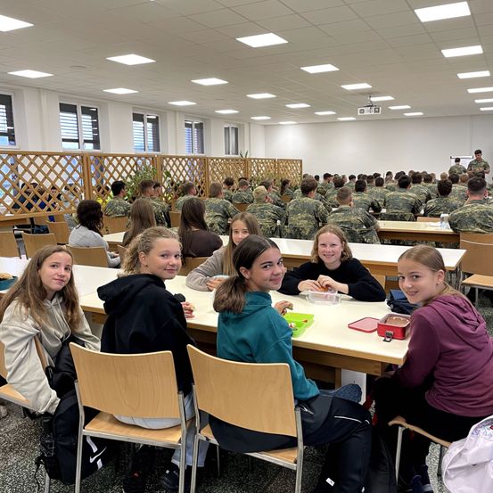 Ein Speisesaal mit Tischen und Stühlen, in dem eine Gruppe junger Mädchen sitzt und lächelt. Im Hintergrund befinden sich Menschen in Militäruniformen. Der Saal hat weiße Wände, eine Decke mit Beleuchtung und Fenster mit Jalousien.