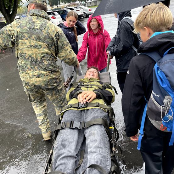Ein Mann wird auf einer Trage von einer Person in Tarnkleidung und einer anderen in einer Regenjacke getragen, während ein Kind einen Regenschirm hält. Ein junger Junge mit Rucksack beobachtet.