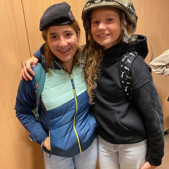 Zwei Mädchen stehen zusammen; eine in einer blauen Jacke, Barett und Rucksack, die andere in einer schwarzen Jacke mit Helm und Rucksack. Sie lächeln für ein Foto in einem Raum mit Holzwänden.