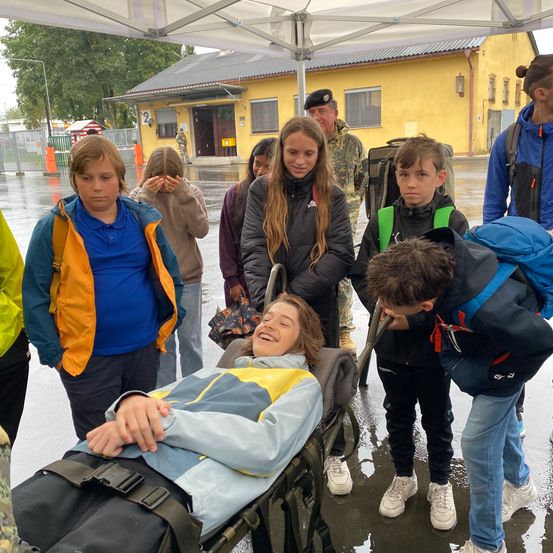 Ein Mädchen im Krankenhausbett lächelt, während eine Gruppe von Kindern bei Regen um sie herum steht, ein Erwachsener steht hinter ihnen.