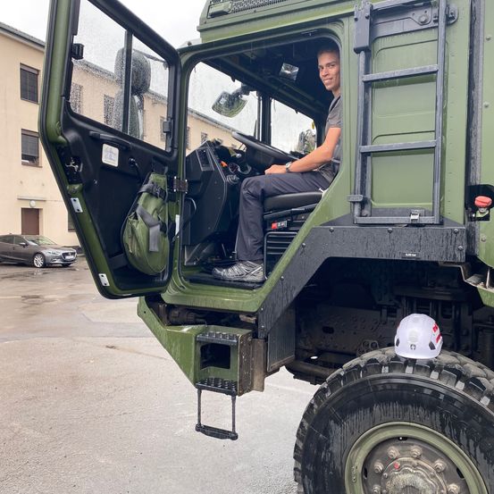 Eine Person sitzt in einem grünen Militärfahrzeug. Ein weißer Helm ist auf der Reifen, und ein Auto ist hinter dem Fahrzeug geparkt.