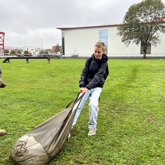 Ein Junge trägt einen großen, abgenutzten Beutel durch ein Grasfeld. Hinter ihm steht ein modernes Gebäude mit Glasfenstern und einem Baum. Mehrere Verkehrskegel sind in einer Reihe aufgestellt. In der Ferne umgibt ein Zaun einen Parkplatz mit mehreren Autos.