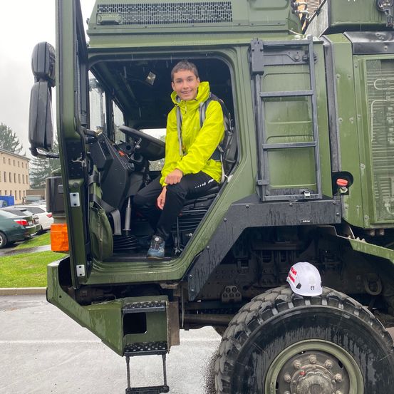 Ein junger Junge sitzt lächelnd in einem großen grünen Militärfahrzeug. Er trägt eine gelbe Regenjacke und Turnschuhe. Ein weißer Harthelm liegt auf dem Reifen unter ihm.