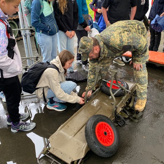 Ein Soldat in Tarnkleidung hilft einem jungen Mädchen beim Reparieren eines Militärfahrzeugs draußen auf einer nassen Oberfläche. Mehrere Zuschauer, darunter Kinder, beobachten.