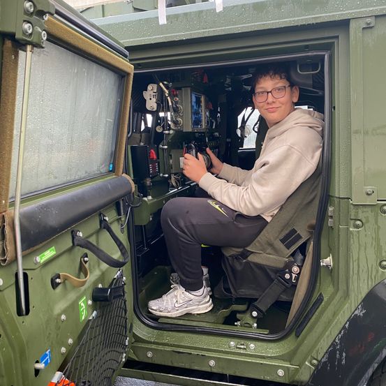 Ein junger Mann sitzt in der offenen Tür eines Militärfahrzeugs, trägt eine Brille und eine beigefarbene Kapuzenjacke. Er hält das Lenkrad mit beiden Händen fest.