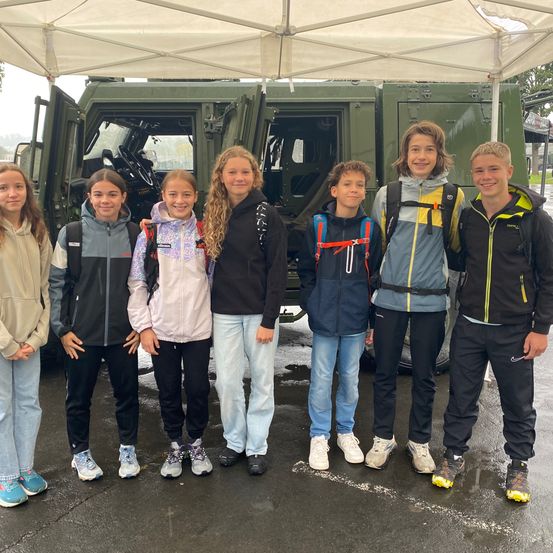Eine Gruppe junger Schüler, einige Mädchen und Jungen, steht draußen neben einem Militärfahrzeug. Sie tragen Jacken und Turnschuhe. Das Wetter scheint regnerisch zu sein.