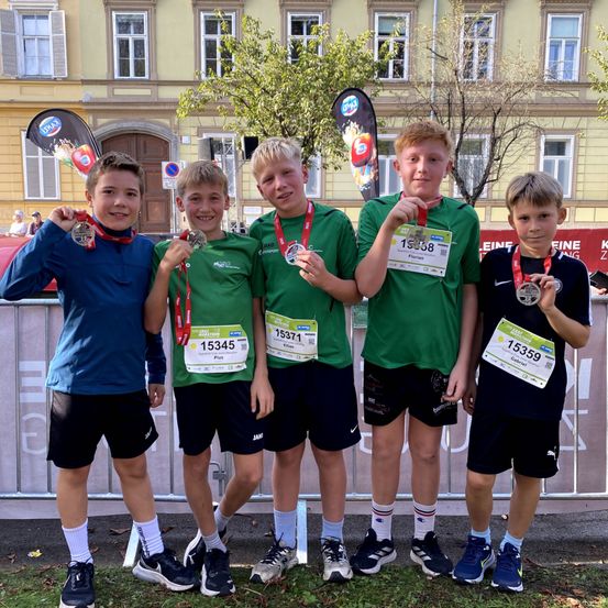 Fünf junge Jungen in grünen und blauen Uniformen stehen zusammen und halten Medaillen. Sie stehen vor einem Gebäude mit vielen Fenstern.