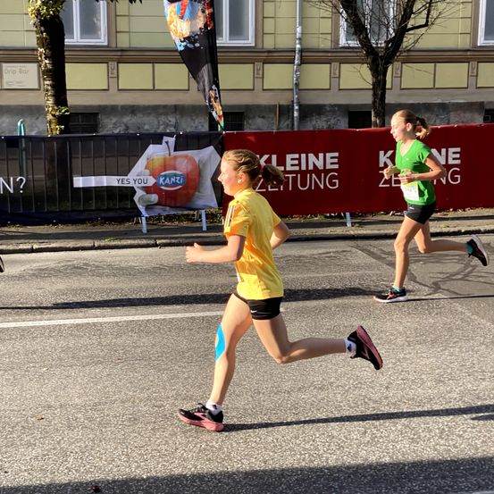 Zwei Mädchen laufen einen Marathon. Das Mädchen im gelben Shirt führt. Dahinter ist ein anderes Mädchen in Grün.