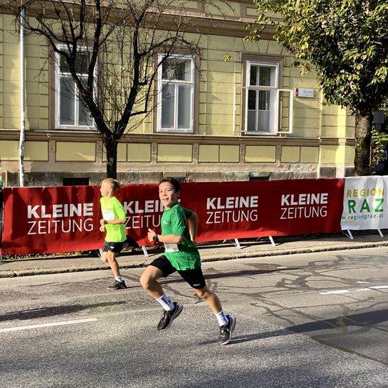 Zwei Läufer sprinten eine Stadtstraße hinunter, hinter ihnen ein Banner für Kleine Zeitung.