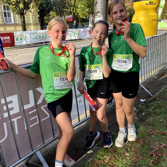 Drei junge Mädchen in grünen Shirts mit Medaillen um den Hals posieren für ein Foto. Sie stehen neben einem Metallzaun, mit einem Grasbereich und Bäumen dahinter.
