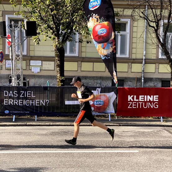 Ein Mann läuft auf der Straße, hinter ihm ein rotes Banner. Das Banner steht für 'Kleine Zeitung'. Ein weiteres Banner mit Apfellogo und Text steht links. Im Hintergrund befinden sich Gebäude, Bäume und Lautsprecher.