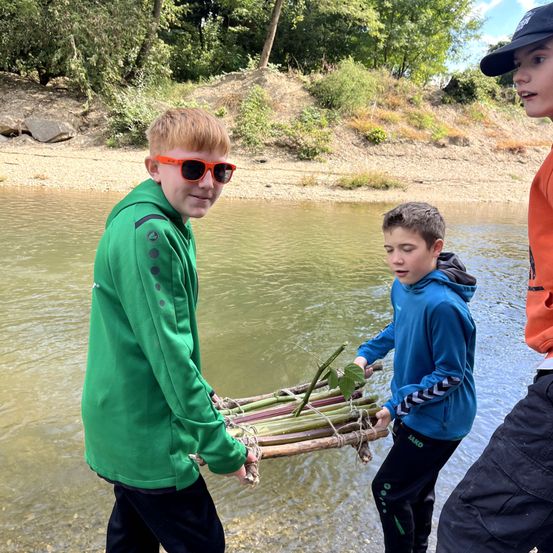 Drei Jungen sind in einem Fluss und tragen ein Bündel Stöcke. Der Junge im grünen Jacken trägt eine Sonnenbrille. Der Junge neben ihm in einer blauen Jacke hält ein Bündel Stöcke. Der dritte Junge trägt ein orangefarbenes Hemd.