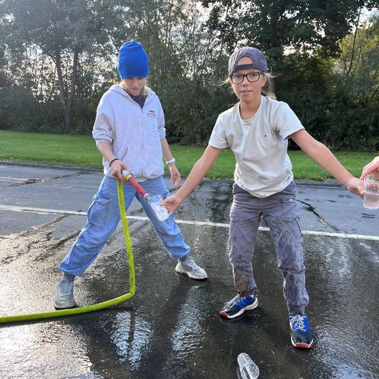 Zwei Mädchen spielen mit einem Wasserschlauch auf einer nassen Straße, eines hält eine Wasserflasche und das andere einen Becher. Sie tragen Turnschuhe und stehen auf nassem Bürgersteig.