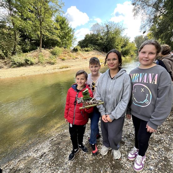 Vier Kinder stehen am Ufer eines Flusses mit Bäumen im Hintergrund. Die Kinder lächeln, eines hält eine Pflanze.