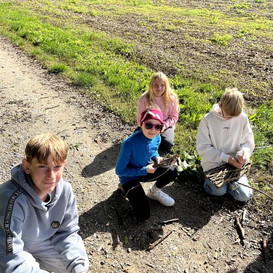 Vier Kinder sitzen auf einem Feldweg in einem Grasfeld. Eines hält einen Bündel Äste.