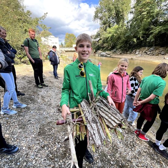 Ein Junge hält ein Bündel Stöcke in der Nähe eines Flusses. Hinter ihm beobachten eine Gruppe von Kindern und Erwachsenen. Der Fluss hat eine Brücke im Hintergrund.