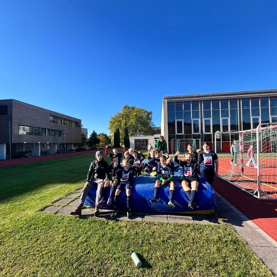 Eine Gruppe junger Athleten posiert für ein Foto auf einem blauen Luftkissen, mit Gebäuden und einem Fußballtor im Hintergrund.