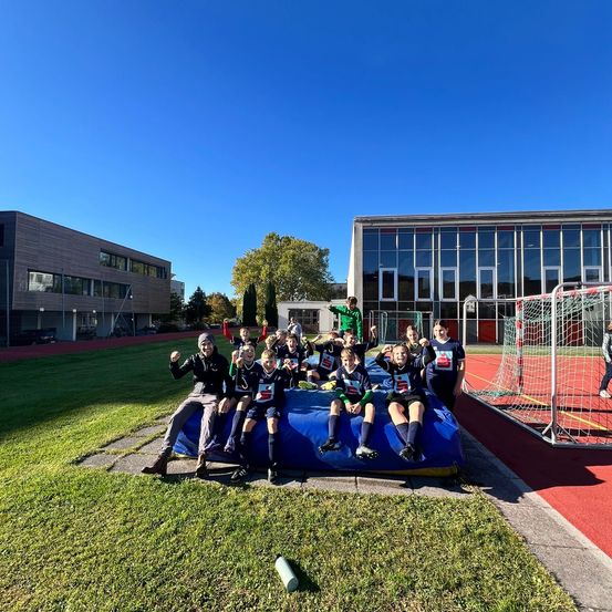 Eine Gruppe von Kindern in Sportuniformen sitzt auf einem blauen aufblasbaren Objekt und posiert für ein Foto. Sie stehen vor einem Gebäude mit Glasfenstern und einem Fußballtor.