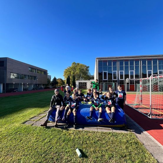 Eine Gruppe von Kindern und zwei Erwachsenen sitzt auf einem blauen Aufblasbaren und posiert für ein Foto. Dahinter befindet sich eine Rasenfläche mit einem Gebäude im Hintergrund. Rechts steht ein Fußballtor.