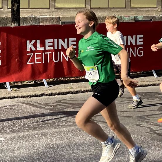 Eine junge Frau in einem grünen Shirt mit der Nummer 556 läuft in einem Marathon mit einem lächelnden Gesicht. Hinter ihr läuft ein Junge.