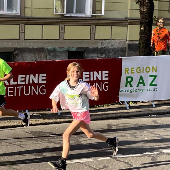 Ein junges Mädchen läuft in einem Marathon, trägt ein grün-weißes Shirt mit der Nummer 15353. Ein anderer Läufer ist hinter ihr. Im Hintergrund befindet sich ein Gebäude mit Fenstern und einem Baum.