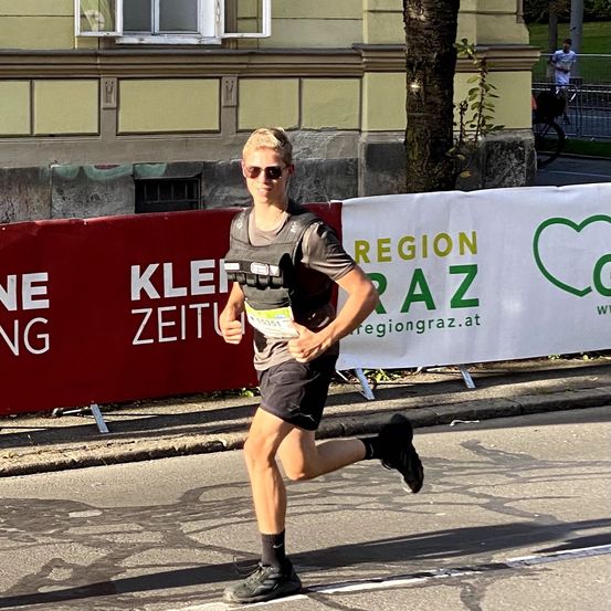 Eine Person läuft auf der Straße und trägt eine Sonnenbrille und ein Laufgeschirr. Hinter ihr befinden sich Banner und ein Gebäude mit Fenstern.