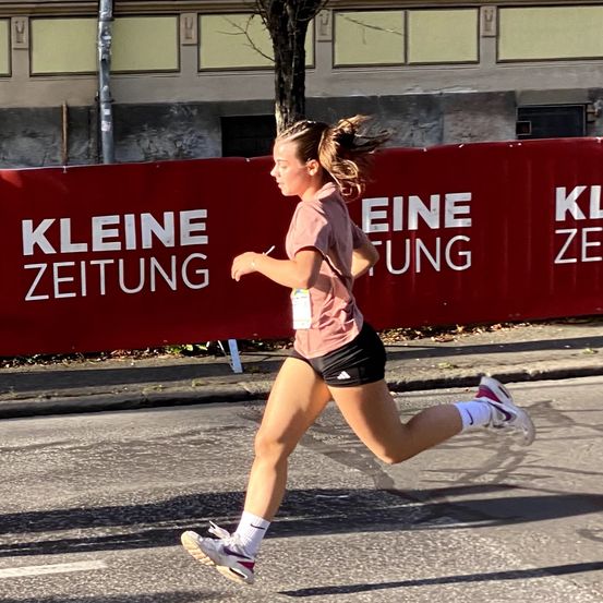 Eine Frau läuft auf einer Stadtstraße vor einem roten Banner mit dem Text 'Kleine Zeitung'. Sie trägt ein rosa Shirt, schwarze Shorts und Turnschuhe.