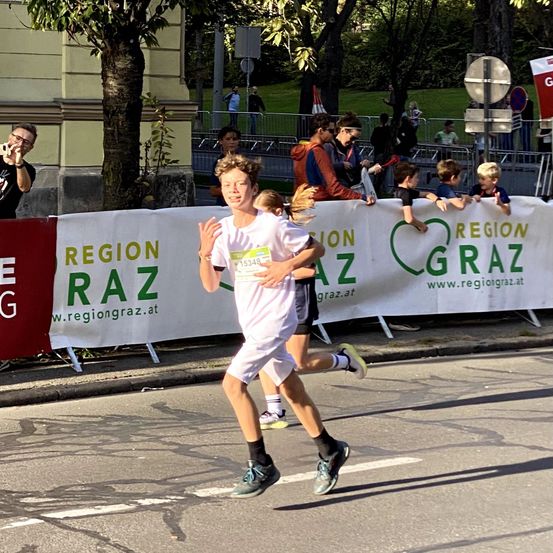 Ein Marathonläufer mit dem weißen Trikot und der Nummer 15348 läuft auf der Straße, mit einem Banner für die Region Graz im Hintergrund.