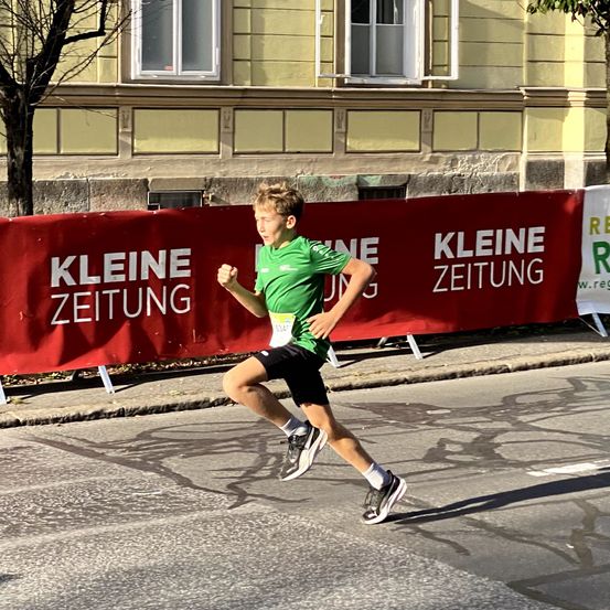 Ein junger Junge läuft einen Marathon, trägt ein grünes Shirt und schwarze Shorts. Er ist neben einem roten Banner und hinter ihm steht ein Gebäude mit Fenstern.