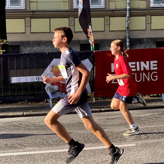 Ein Junge und ein Mädchen laufen einen Marathon. Sie tragen Sportkleidung und Schuhe. Der Junge hat einen Nummernanhänger an seinem Shirt. Hinter ihnen ist ein roter Banner mit weißen Buchstaben.
