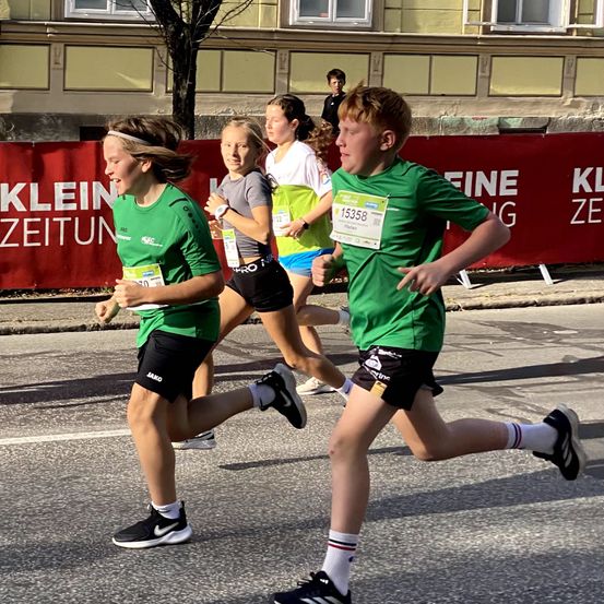 Vier Läufer in grünen und grauen Trikots nehmen an einem Marathon in einem städtischen Umfeld teil, mit Gebäuden und einem Banner im Hintergrund.