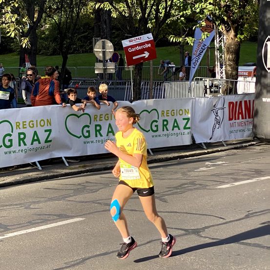 Eine Läuferin in einem gelben Shirt mit einem Startnummernband mit der Nummer 15845 läuft auf einer Straße mit einem Banner, das Region Graz sagt. Hinter ihr schauen Leute von hinter einem Zaun zu. Bäume und ein Schild mit einem Pfeil sind im Hintergrund sichtbar.