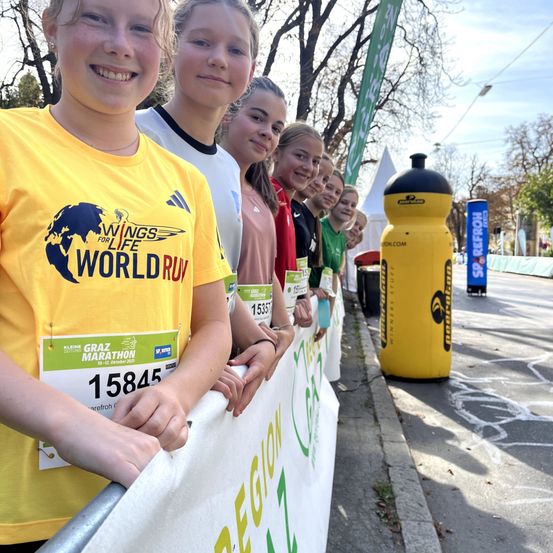 Eine Gruppe junger Läufer, die passende gelbe T-Shirts tragen, posiert für ein Foto am Straßenrand. Das Shirt trägt den Schriftzug 'Wings for Life World Run'. Eine gelbe Wasserflasche befindet sich in der Nähe.