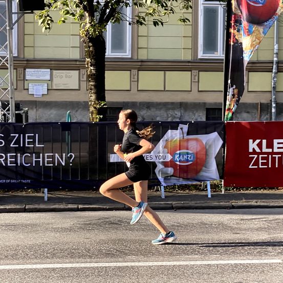 Eine junge Frau läuft die Straße hinunter vor einem Gebäude mit Schildern, die 'Is Ziel Erreichen?' und 'Kle Zeit' sagen.
