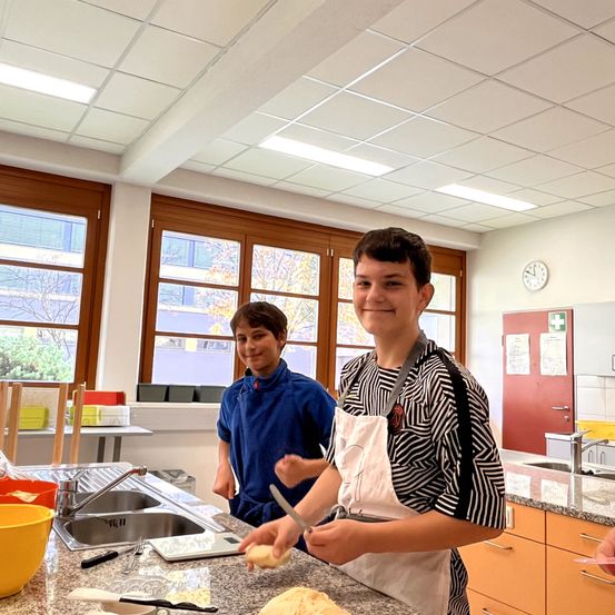 Zwei Jungen in einer Küche, einer in einem blauen Hemd und der andere in einer weißen Schürze, bereiten Essen zu. Sie stehen vor einem Waschbecken mit Wasserhahn, und verschiedene Küchenutensilien sind um sie herum. Hinter ihnen befinden sich Fenster mit Blick auf Bäume und Gebäude.