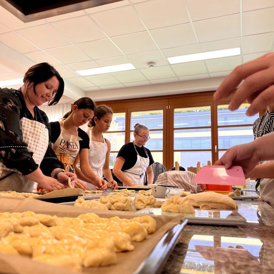 Eine Gruppe von Frauen befindet sich in einer Küche und bereitet Teig auf Blechen zu. Eine Frau trägt eine Schürze mit dem Wort Pasta. Der Teig ist auf Backblechen und einige bereits auf dem Tisch.