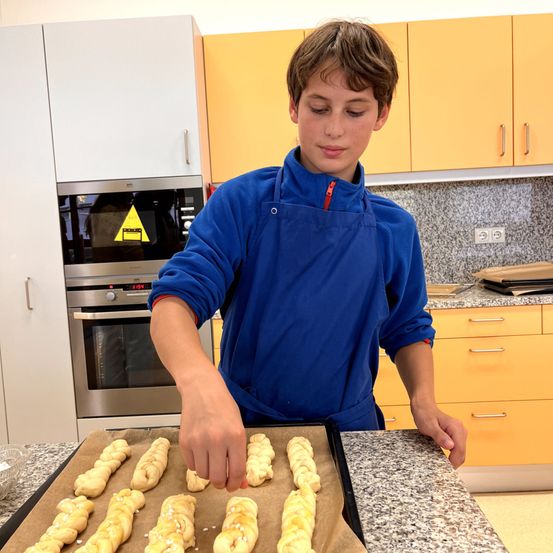 Ein junger Junge in einem blauen Mantel macht Brot in der Küche. Er legt den Teig auf ein Backblech. Hinter ihm befindet sich ein Mikrowellenherd und eine Arbeitsplatte mit Schränken.