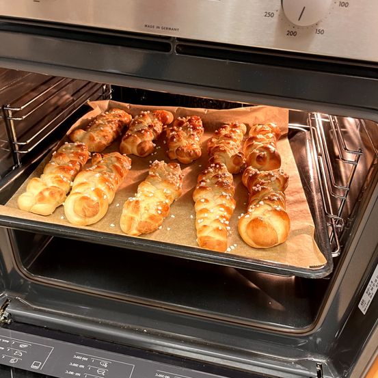 Ein Backblech mit mehreren geflochtenen Brötchen ist in einem Ofen. Die Brötchen sind goldbraun und mit Sesam bestreut. Der Ofen ist offen, und die Temperatur ist auf 200 Grad eingestellt.