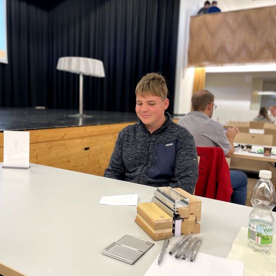Ein Junge sitzt an einem Tisch mit einer Holzkiste und Stiften und lächelt. Hinter ihm sitzt eine weitere Person in einem Stuhl. Eine Projektionsleinwand befindet sich links und eine Flasche Wasser steht auf dem Tisch.