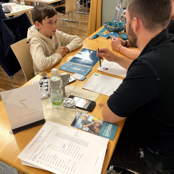Ein Mann sitzt mit einem Jungen an einem Tisch, beide schauen nach vorne. Auf dem Tisch liegen Papiere, ein Handy und eine Flasche. Der Junge trägt eine weiße Kapuzenjacke, während der Mann einen Bart hat. Hinter ihnen stehen Stühle und ein Tisch mit einer blauen Schüssel und anderen Gegenständen.