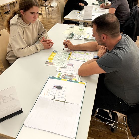 Eine Frau und ein Mann sitzen an einem Tisch und überprüfen Dokumente. Die Frau hat die Hände gefaltet. Der Mann hält einen Stift und konzentriert sich auf die Papiere.