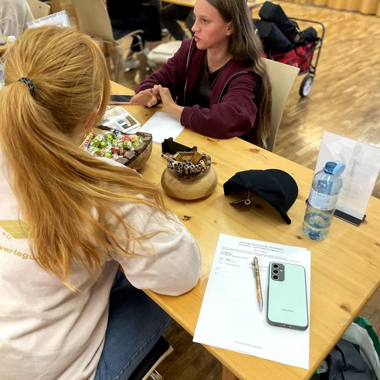Zwei Frauen sitzen an einem Tisch, eine hält ein Telefon und die andere hat eine Schüssel mit Süßigkeiten. Ein Telefon, ein Stift und eine Wasserflasche auf dem Tisch. Eine Person mit einem Trolley im Hintergrund.