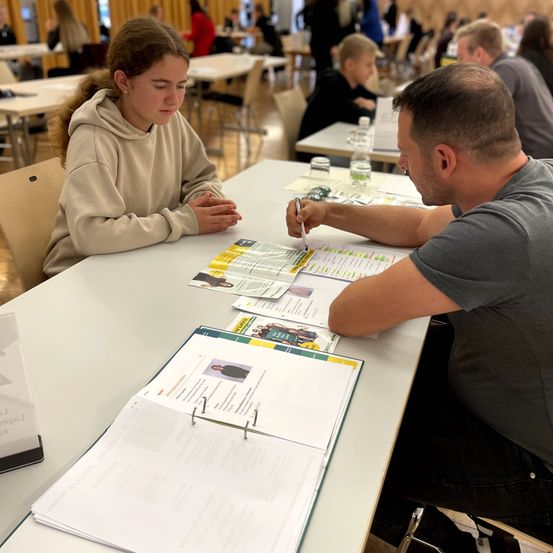 Ein Mann und ein Mädchen sitzen an einem Tisch und besprechen möglicherweise ein Dokument. Sie halten beide Stifte in der Hand. Der Mann scheint dem Mädchen etwas zu erklären.