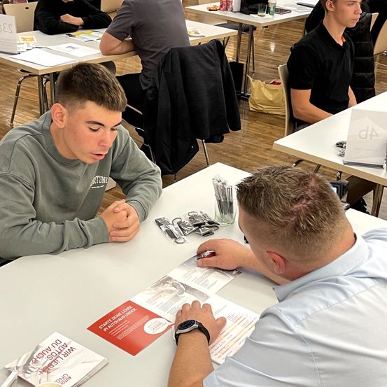 Eine Gruppe von Personen in einem Klassenzimmer, wobei ein Mann in Grau mit einem anderen Mann in Hellblau interagiert. Sie sitzen an einem Tisch mit verschiedenen Gegenständen darauf.