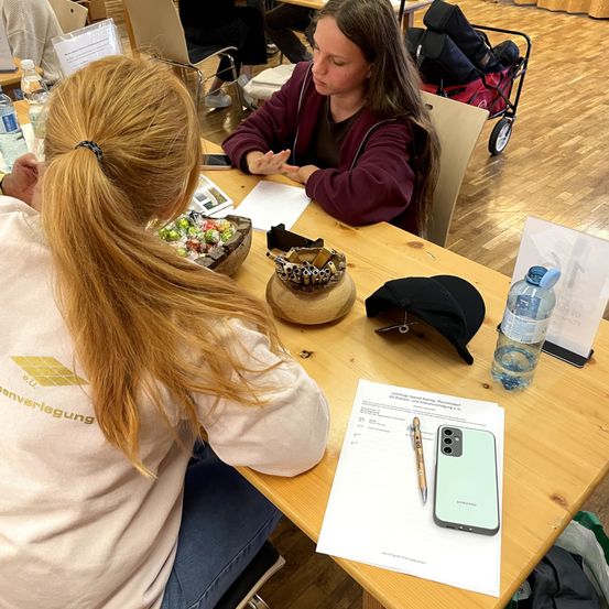 Zwei Frauen sitzen an einem Holztisch mit verschiedenen Gegenständen. Eine Frau hat eine Schüssel mit Süßigkeiten, während die andere ein Handy und Dokumente hat. Ein Samsung-Handy, eine Wasserflasche und eine schwarze Kappe stehen auf dem Tisch. Der Raum hat Holzböden.