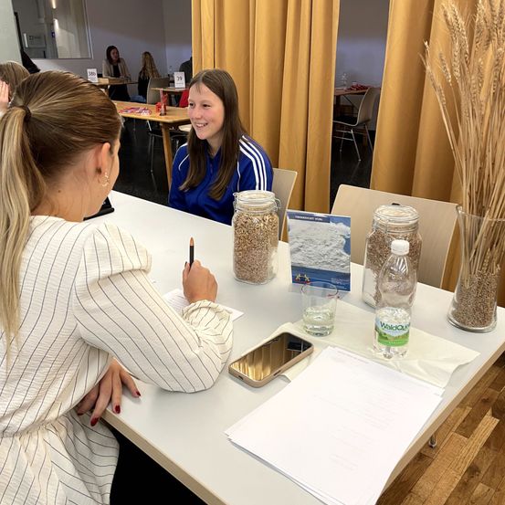 Zwei Frauen sitzen an einem Tisch, eine in einem blauen Mantel schreibt lächelnd. Sie haben Gläser mit Getreide, eine Wasserflasche und ein Handy auf dem Tisch. Hinter ihnen sind gelbe Vorhänge und ein Stuhl zu sehen.