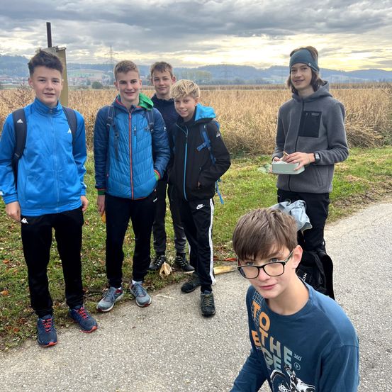 Fünf Jungen stehen auf einem Weg in einem Grasfeld. Sie tragen Jacken und Sneakers, und einer hält eine Box. Hinter ihnen steht ein Pfosten im Feld.