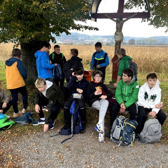 Schüler sitzen auf einer Bank, einige essen, unter einem Baum mit einem Kruzifix. Sie tragen Jacken und Turnschuhe.